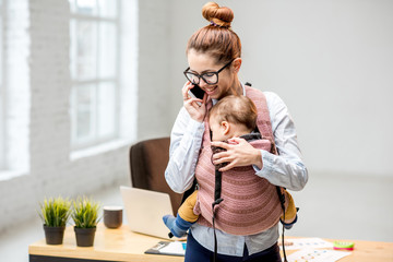 Young multitasking businesswoman standing with her baby son during the work at the office