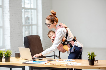 Young multitasking businesswoman standing with her baby son during the work at the office