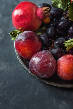 Organic Purple Grapes Red Plums Pomegranates on Vintage Plate. Black Stone Background. Autumn Fall Still Life Harvest Jewish New Year Rosh Hashanah Thanksgiving Copy Space