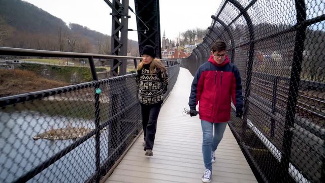 Steadicam gimbal shot of pretty mature blonde woman and teenage son walk across a foot bridge next to a railroad track in Harpers Ferry National Park.