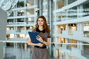 The young business woman in gray office dress holds in hand folders with documents. The girl costs against the background of office premises of business center.