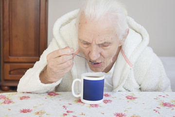 Stara kobieta w szlafroku siedzi przy stole i pije herbatę z łyżeczki.