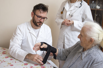 Pielęgniarz w białym kitlu zakłada stabilizator na nadgarstek starej kobiety przy nadzorze lekarza. - obrazy, fototapety, plakaty