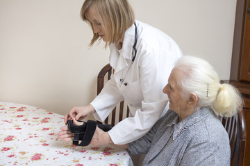 Lekarka zakłada stabilizator na rękę starej kobiety siedzącej przy stole.