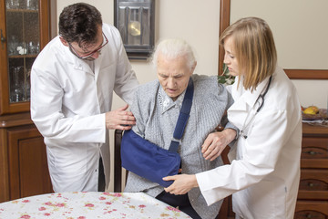 Lekarka i pielęgniarz pomagają wstać starej kobiecie z krzesła. Staruszka z ręką na temblaku. - obrazy, fototapety, plakaty