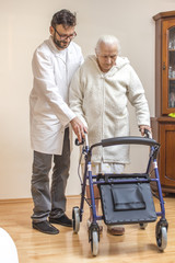 Staruszka w białym szlafroku chodzi przy pomocy chodzika przy asyście pielęgniarza.