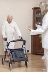 Staruszka w białym szlafroku uczy się chodzić z balkonikiem przy asyście pielęgniarki.