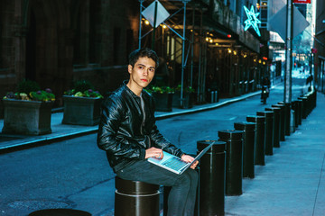 Asian American college student studying in New York. Wearing black leather jacket, pants, a young guy sitting on narrow street, reading, working on laptop computer, thinking. Filtered look..