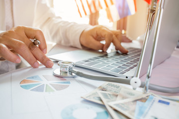 Doctor working on laptop computer with report analysis and money about Healthcare costs and fees in medical hostpital office. Focus stethoscope on table. Healthcare budget and business concept