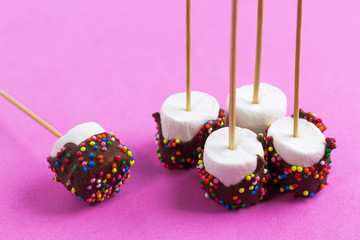 marshmallows on a sticks, laying on pink background. glazed with chocolate and colorful sprinkles.