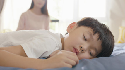 Fototapeta na wymiar Asian boy sleeping in bed in the morning while mother sitting in background