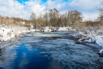 the blue river is covered with a thin transparent layer of ice, on the banks and trees there is white snow, a winter landscape