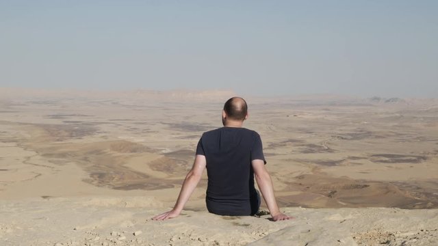 Back view of man seated on the edge, admiring beautiful landscape of the Makhtesh Ramon Crater, Israel.
