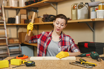 Pretty caucasian young brown-hair woman in plaid shirt, gray T-shirt, yellow gloves working in carpentry workshop at wooden table place with different tools, hammering nails into board with hammer.
