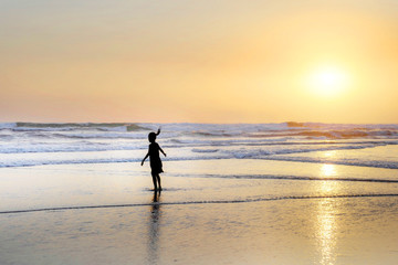 Silhouette of anonymous female child running and playing on amazing beautiful desert beach on sunset with an orange sky and golden light