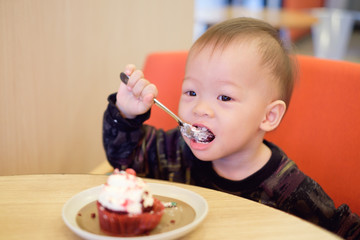 Cute Happy Smiling little Asian 18 months / 1 year old toddler baby boy child eating cupcake with fork, he's sitting on chair at restaurant, Children & Self feeding concept, Soft & Selective focus