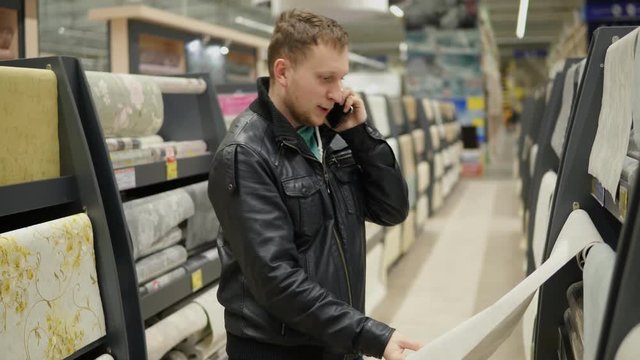 Young man is calling someone to be consulted while choosing wallpapers in supermarket. He is looking at the wallpaper, touching it and thinking which one is better