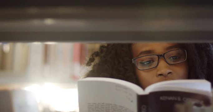 Close up of the African American young curly woman student in glasses reading a book among books shelves in the library. Portrait. Inside
