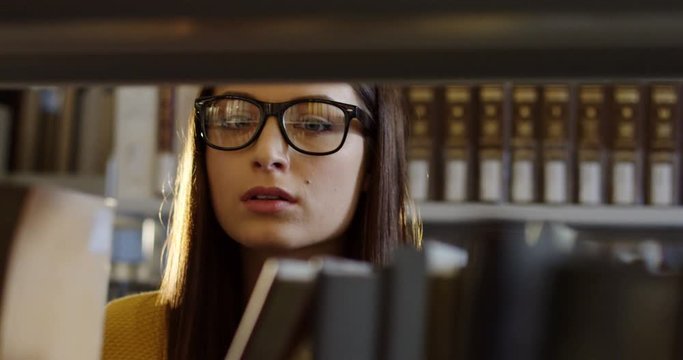 Close up of the young attractive woman student in glasses chosing a book among books on the shelves in the library. Portrait shot. Indoors