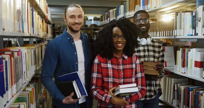 Portrait of three African American and Caucasian male and female students standing in the library among books shelves and holding books in their hands. Indoor