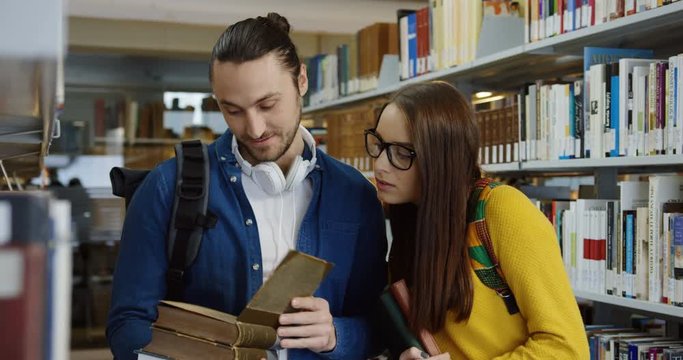 Male and female Caucasian students standing in a library passage and flipping an old book from a shelf. Indoors. Portrait shot