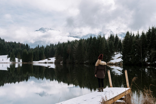 Young female standing at Lake Geroldsee overlooking a forest with cabin with the Karwendel Mountains in Background in Bavaria, Germany in winter