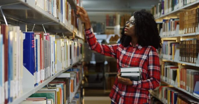 Portrait of the young pretty African American woman in glasses looking for a book on the books shelves in the library. Indoors