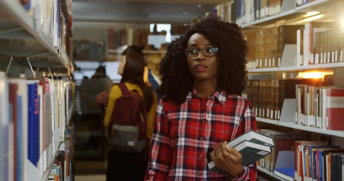 Close up of the young African American woman in glasses and motley walking in the library and looking for a book from a shelf. Indoor