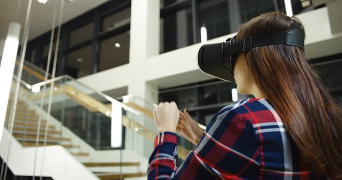 Rear of the young woman with long hair and wearing in plaid shirt having a VR headset, scrolling and taping in the air. Big modern stairs on the background. Indoors