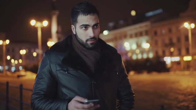 Handsome young man walking down the night snowy street and using his smartphone. Having fun, modern technologies. Slow motion, camera stabilizer shot, male portrait