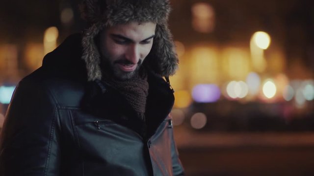 Smiling bearded man in leather jacket and stylish fur hat stands in the city center and uses his smartphone, types messages or surfs the internet. Being online, social networks, modern communication