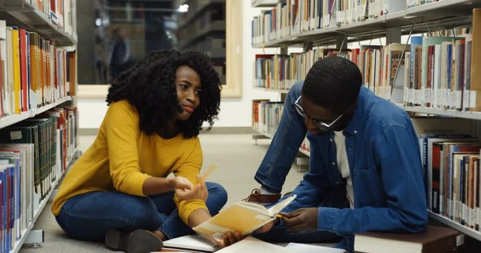 African american young male and female students studying together while sitting on the floor among books shelves at the library passage. Indoor