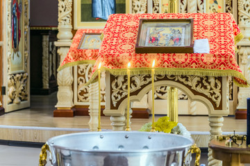 Holy water font for christening ceremony in the Orthodox Church. Three burning candles above the font and a pulpit with an icon for liturgy, prayers and sermons. Selective focus.