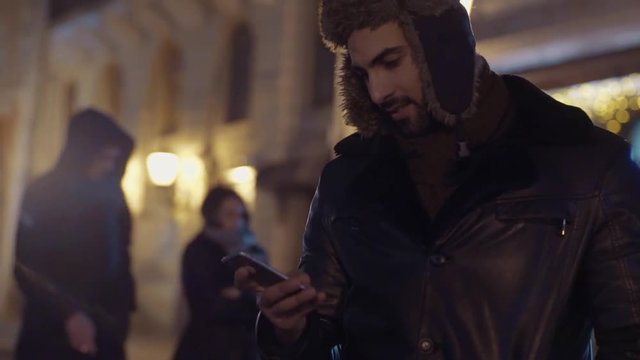 Rotation view of young guy winter wear surfing internet or texting messages via smartphone while standing in the city center. Modern lifestyle, night city lights Social networks, being online