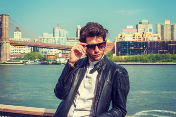 European businessman traveling in New York. Wearing leather jacket, holding sunglasses, a guy with beard, standing by river, looking down, sad, think. Brooklyn bridge, high buildings on background..