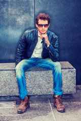 Man Casual Street Fashion. Wearing black leather jacket, white undershirt,  blue jeans, brown boot shoes, sunglasses, a young guy with beard sitting on marble bench against wall, relaxing, thinking. .