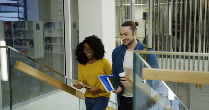 Young multi ethnic man and woman students walking up the stairs and talking cheerfully in the modern library hall. Indoors