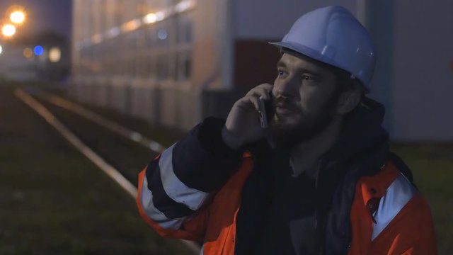 Young and happy worker in hard helmet talking by phone at night. 4k
