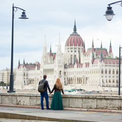Fototapeta premium Parliament Building in Budapest, Hungary. A back view of a couple of tourists holding hands and enjoying the view of the magnificent architecture of Budapest between two ancient lanterns