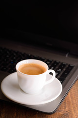 Cup of coffee on a laptop keyboard. Work place modern. Laptop (notebook) with cup of coffee and notepad with pen on old wooden table.