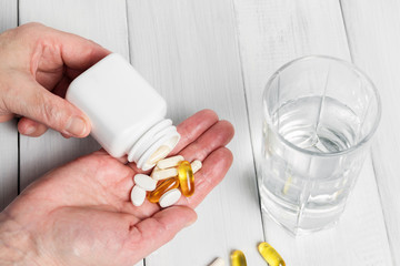 Woman hands hold vitamin complex and dietary supplement, pours from white bottle into palm yellow capsules of omega 3, white pills of calcium and glucosamine, glass of water on wooden planks table