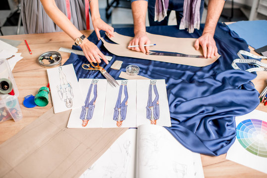 Cutting blue fabric on the table full of tailoring tools. Close-up view on the hands, fabric and fashion drawings