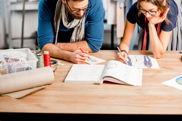 Couple of fashion designers working with clothing sketches at the studio full of tailoring tools and clothes