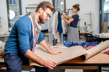 Obraz premium Portret przystojnego projektanta mody siedzącego z papierowymi szkicami w pracowni pełnej narzędzi krawieckich i ubrań