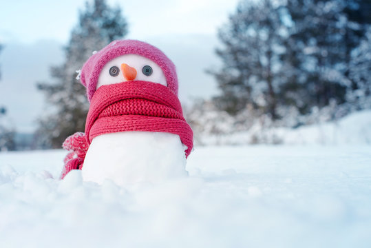 Little snowman girl in a pink knitted hat and a scarf on snow in the winter. Festive background with a lovely snowman. Christmas card, copy space
