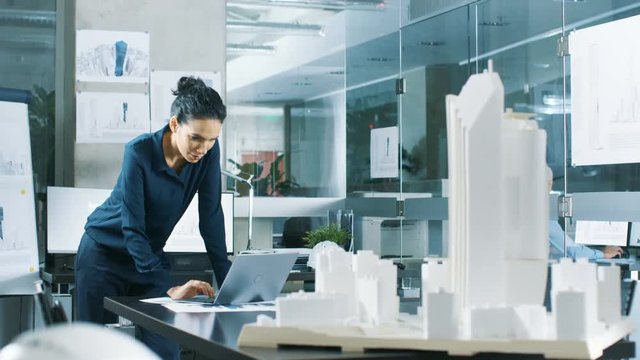 Female Architectural Designer Works on a Laptop,  Engineering New Building Model for the Urban Planning Project. Clean Minimalistic Office, Concrete Walls Covered by Blueprints and Documents. 