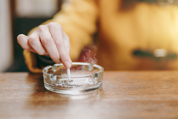 Focus on caucasian young woman hand putting out cigarette on glass ashtray on wooden table,...