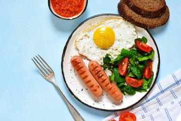 Homemade breakfast: egg, grilled sausage, vegetables, salad on a blue, concrete background.