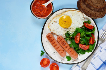Homemade breakfast: egg, grilled sausage, vegetables, salad on a blue, concrete background.