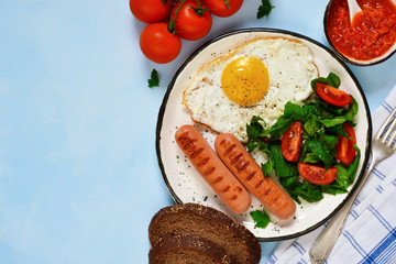 Homemade breakfast: egg, grilled sausage, vegetables, salad on a blue, concrete background.